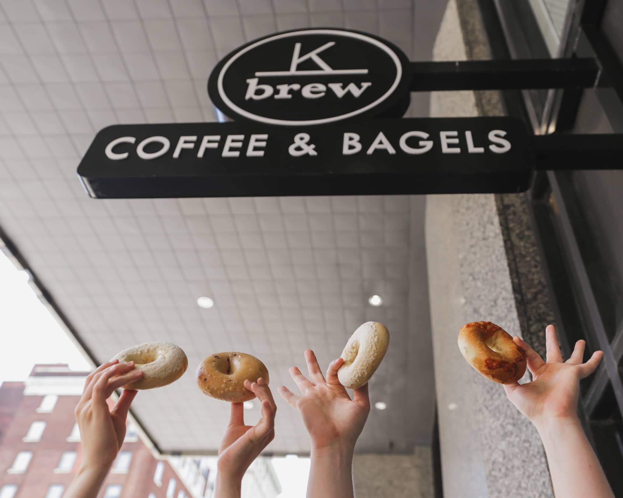 three hands holding up bagels under K Brew's downtown sign
